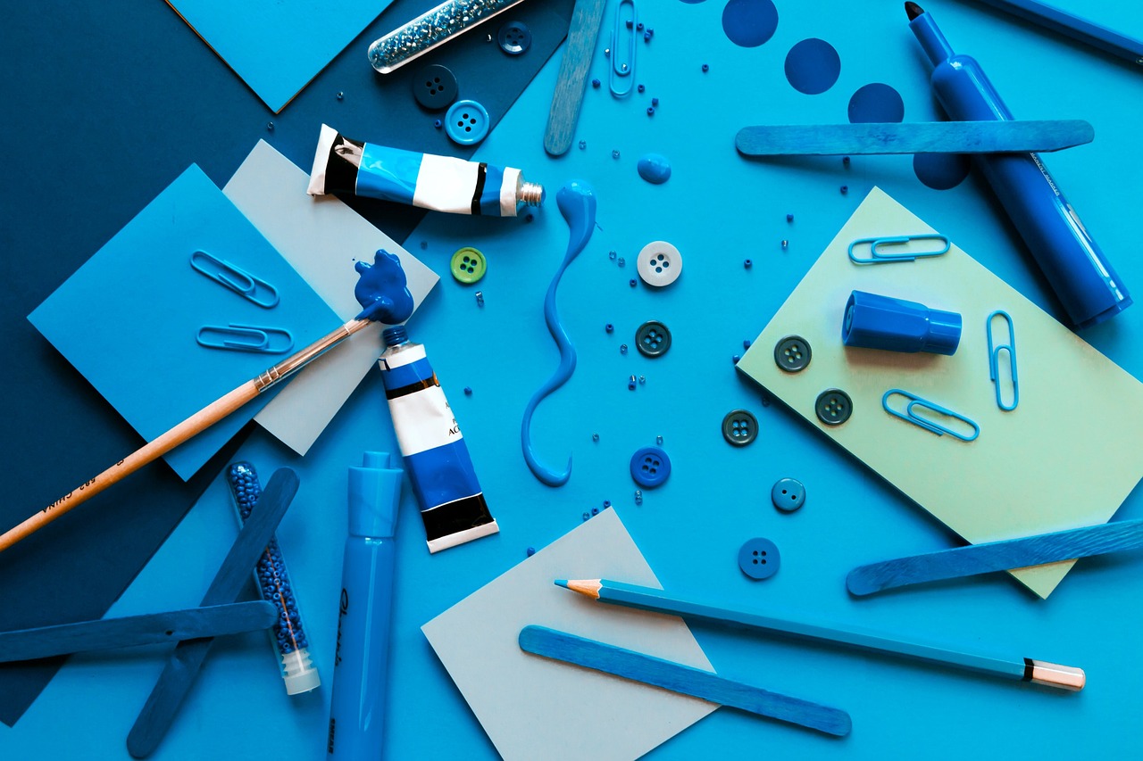 Flat lay of various blue-colored graphic design tools including paint tubes, paper, buttons, pencils, and markers arranged artistically.