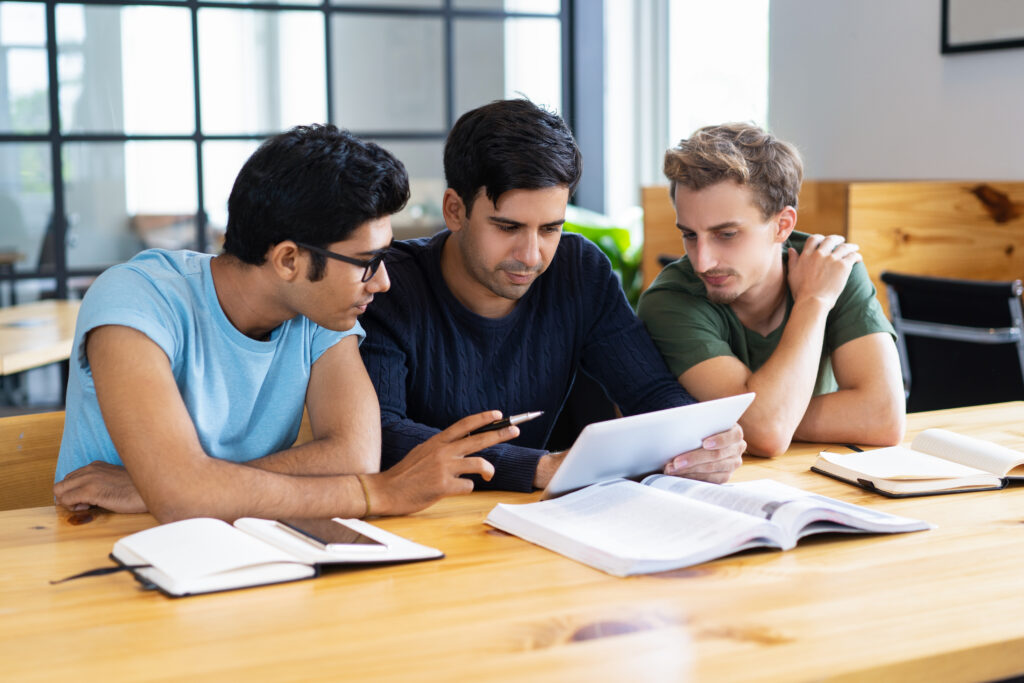 Three young students collaborating at a table with books, notebooks, and a tablet during a group study session.