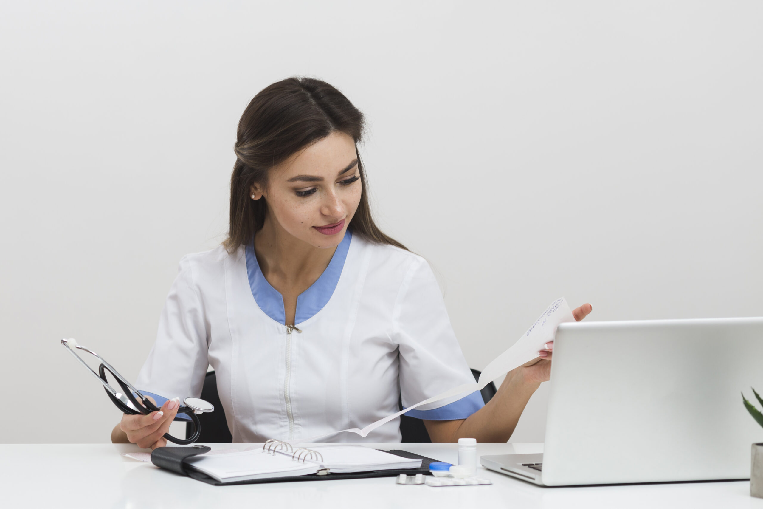 Lady in hospital uniform sitting in front of a laptop with a stethoscope in hand and an open file on the table — representing practical learning in hospital administration.